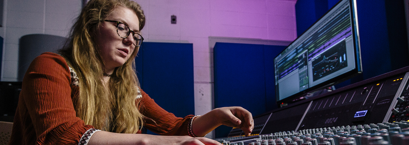 A young woman adjusts the dials on a sound mixing board, a computer screen in the background running sound editing software.