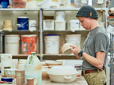 Student surrounded by ceramic creations currently regarding one of them in hand.