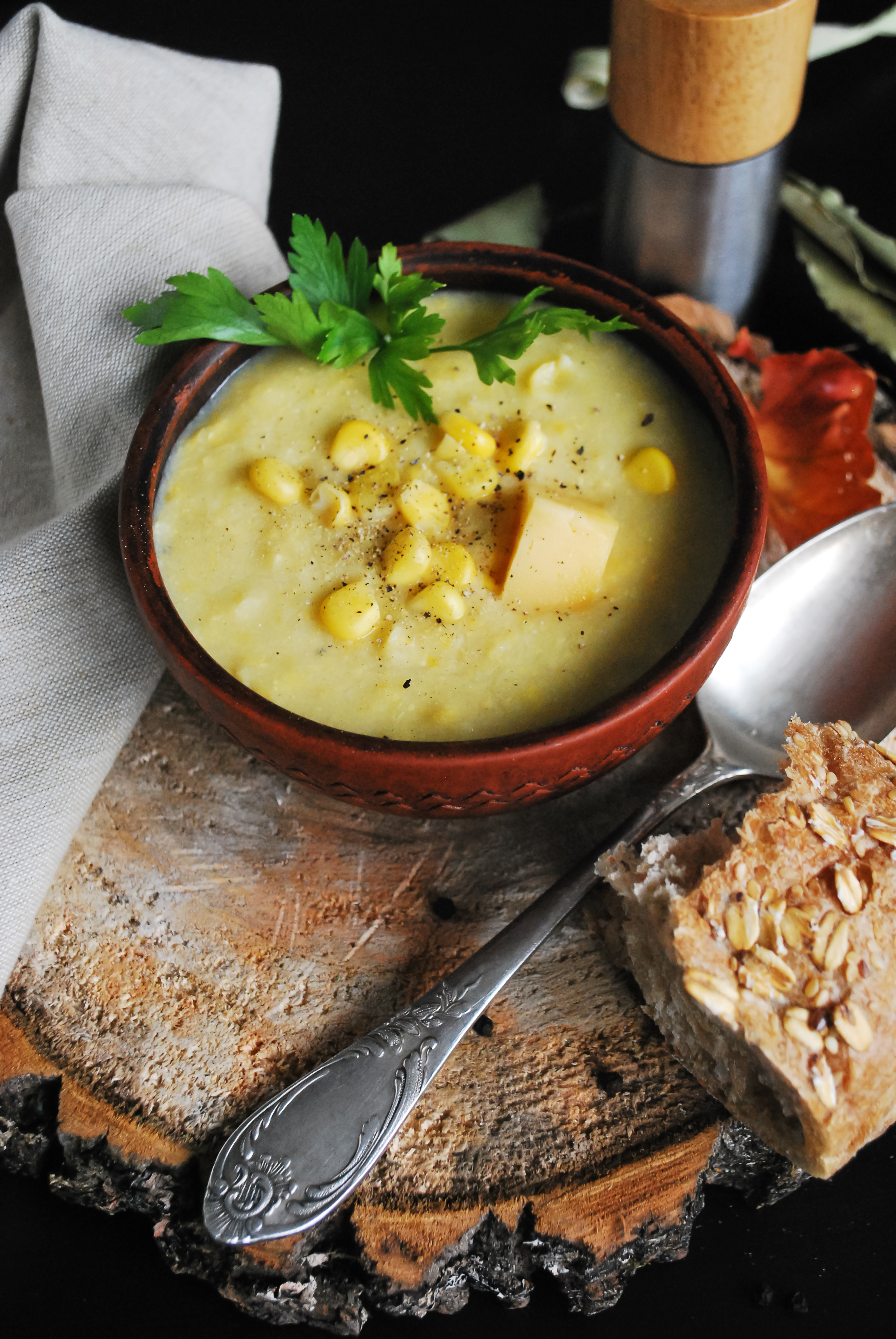 A bowl of potato, corn, and cheese chowder next to a spoon and a piece of bread.