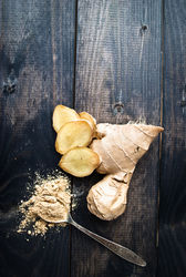 A brown ginger root with chopped pieces and a spoon of ground ginger against a wooden surface.