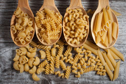 Four wooden spoons containing different shapes of dry pasta noodles.