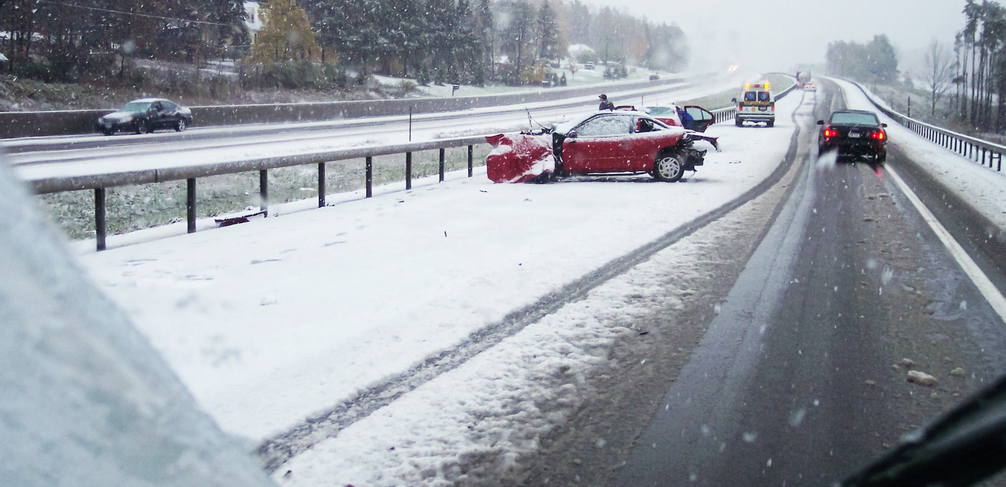 A red car sits on the side of the road, smashed from a car accident. The road is covered in snow and an ambulence sits off in the distance.