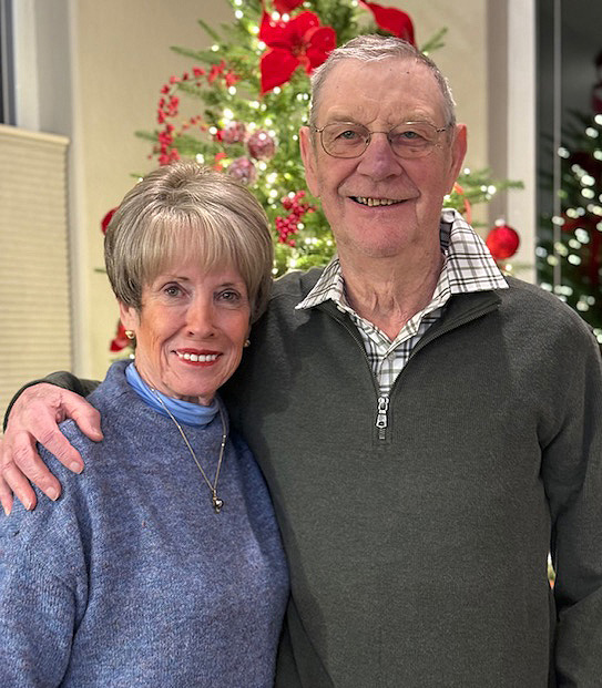 A man and a woman pose for a photo in front of a christmas tree. They are older in age and the man has his arm around the woman.