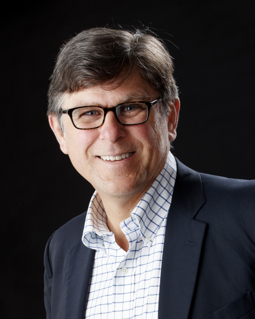 The image shows a man with short, dark hair and glasses, smiling while looking at the camera. He is wearing a dark blazer over a white shirt with a blue checkered pattern. The background is black.