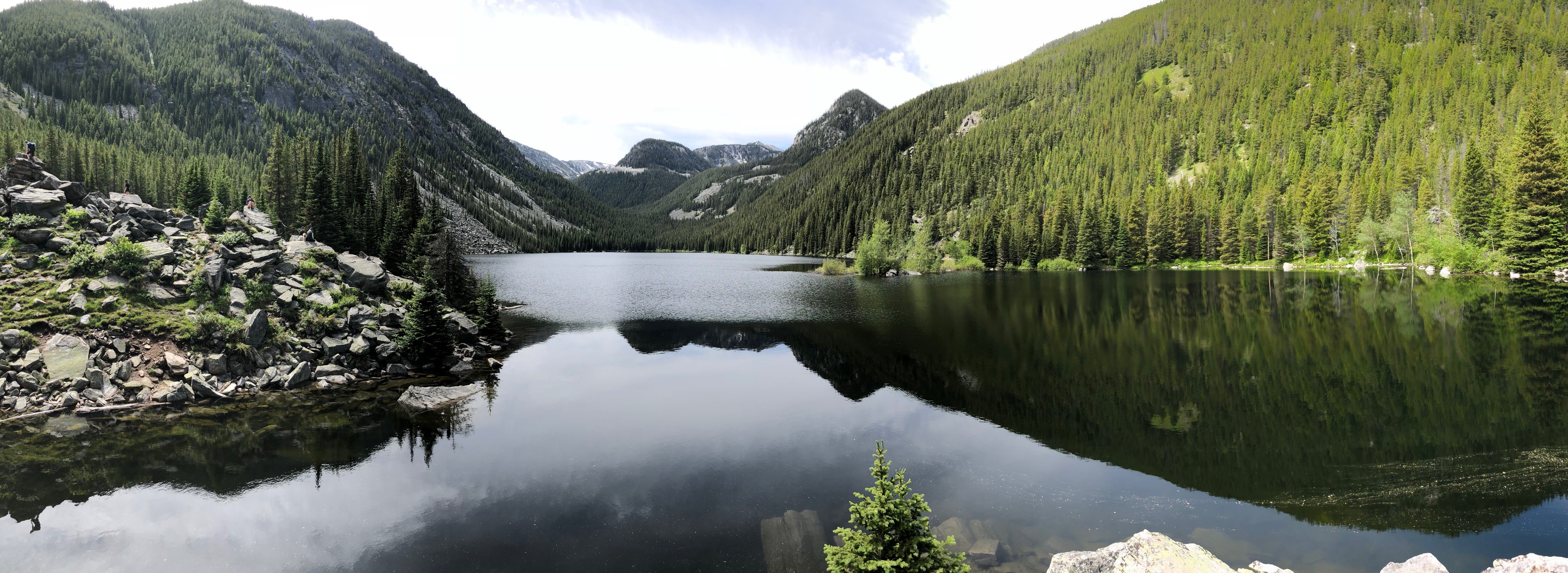  Panoramic view of a serene mountain lake surrounded by lush green forests. The lake reflects the surrounding landscape, creating a mirror image on its surface. Large rocks are visible on the left side of the lake’s shore, and the forested mountains enclose the lake in a tranquil embrace.